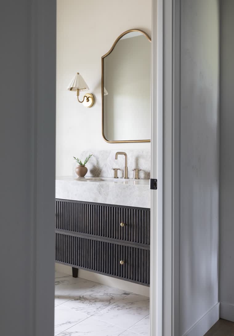 Powder room seen through a doorway, with a fluted dark vanity and a brass mirror