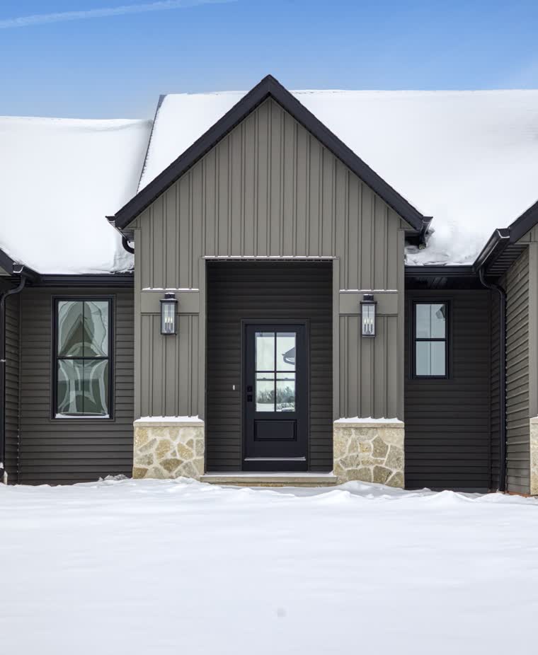 Front entry of a dark board-and-batten home with a stone base, in winter